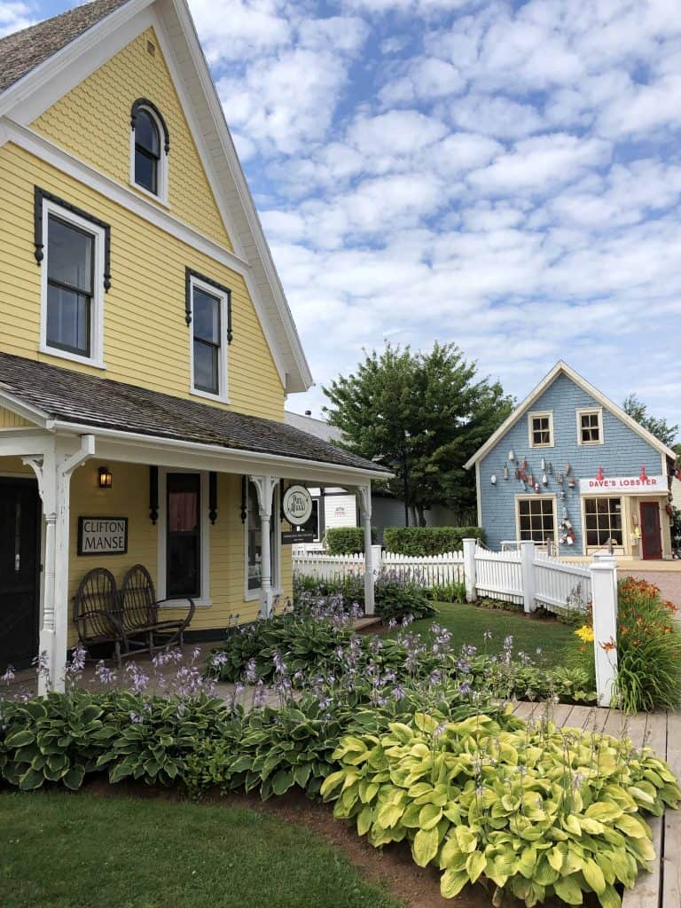 Historic buildings at Avonlea Village in cavendish, Prince Edward Island - yellow building with flowers in front and white picket fence and blue Dave's Lobster building in background.