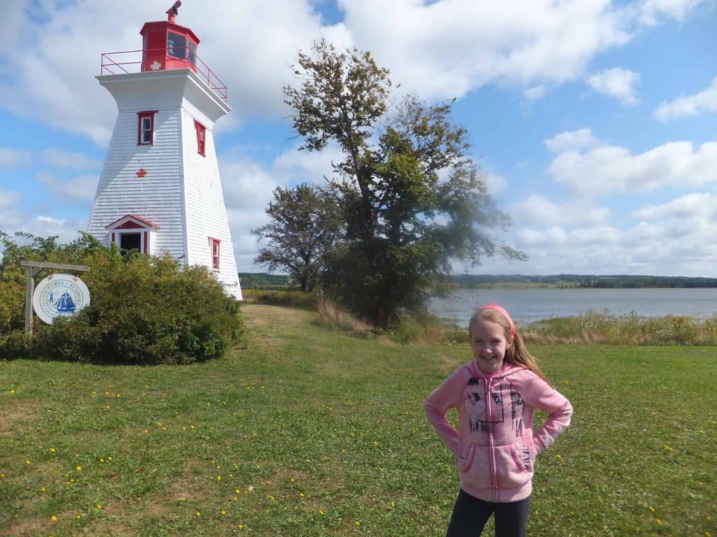 Young girl wearing pink sweatshirt in front of White and red trimmed lighthouse in Victoria-by-the-Sea in Prince Edward Island.