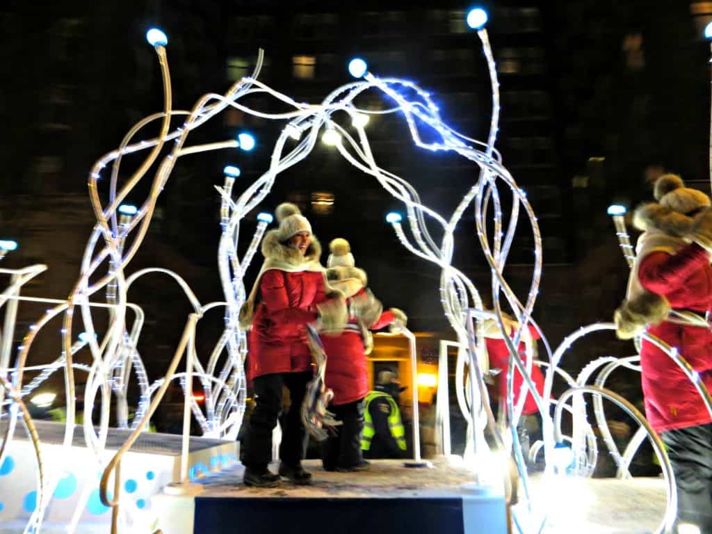 Women in red winter coats, black pants and toques on float surrounded by lights at quebec carnival night parade in quebec City.