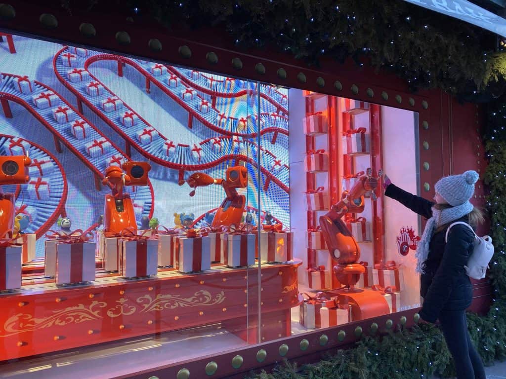 Young woman in dark hat, gray hat and scarf looking at red and blue holiday window display in Toronto.