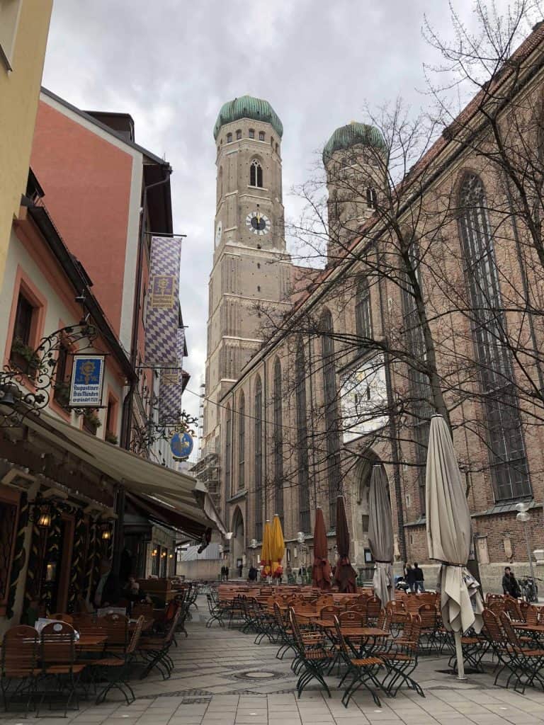 Tables and chairs sitting outside restaurant on street in Munich, Germany with Frauenkirche domed towers in background.