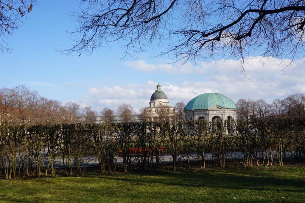 Blue sky with white fluffy clouds, two domed buildings with a row of small bushes without leaves in front in Hofgarten, Munich, Germany.