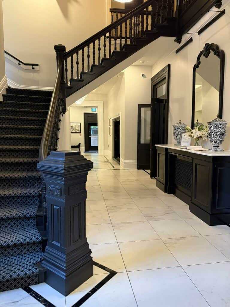 Entrance to The Frontenac Club in Kingston, Ontario - staircase to upper floors, large desk with blue and white vases, black door frames and white and black tiled floor.
