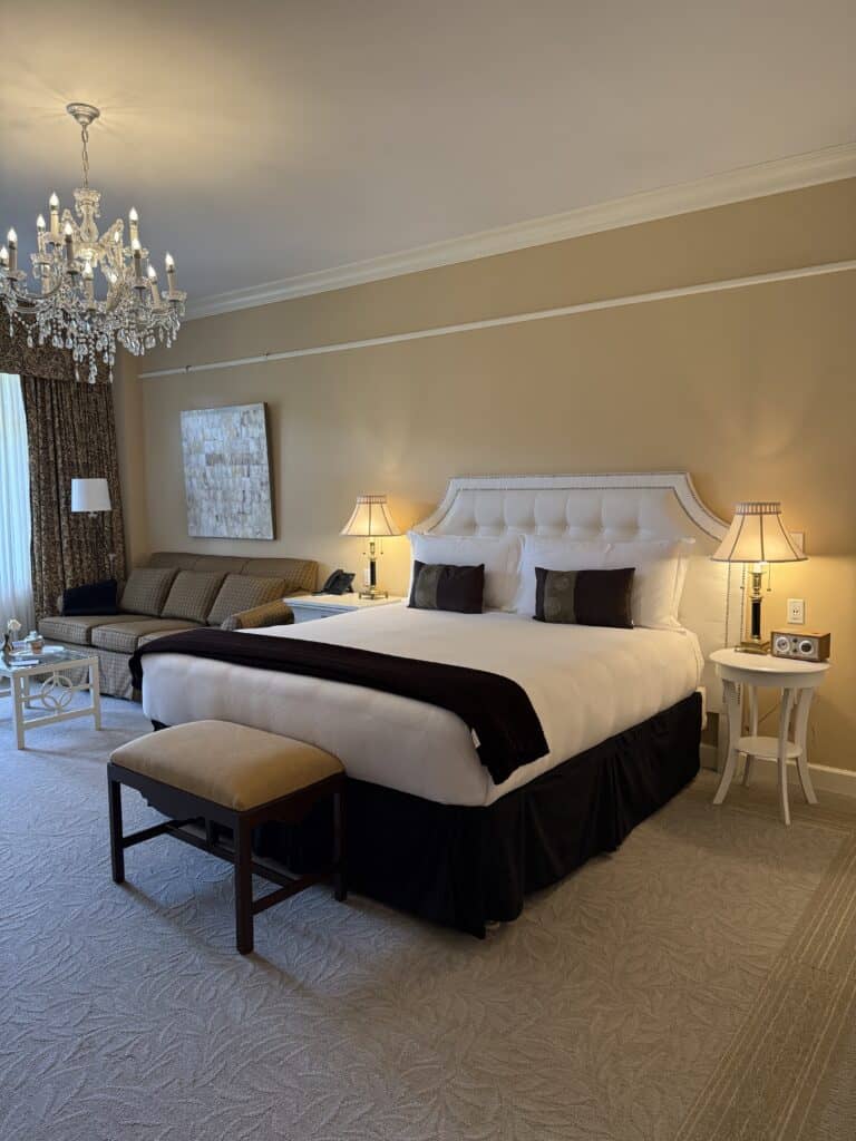 room at the bruce hotel in Stratford, Ontario, glass chandelier, taupe coloured walls, sitting area with couch and bed with upholstered white headboard, white and dark linens and two white bedside tables with lamps.