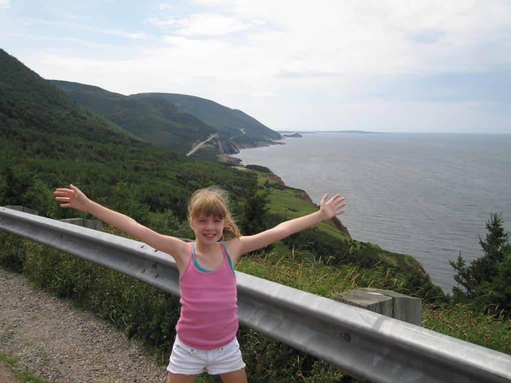 Young girl in white shirts and pink tank top with arms outstretched on roadside along Cabot Trail on Cape Breton Island, Nova Scotia with cliffs and ocean in background.