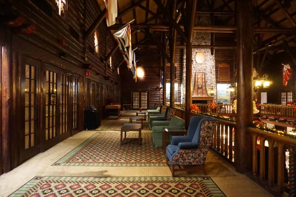 Interior of Chateau Montebello seating area in long hallway with upholstered chairs and tables on carpet along railway with stone fireplace visible in background.