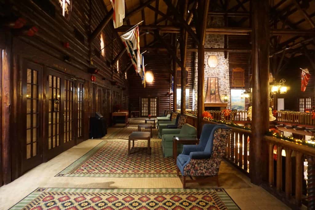 Second floor of Chateau Montebello in Montebello, Quebec - upholstered chairs by wooden railing, patterned rugs on floor with large stone fireplace visible in background.