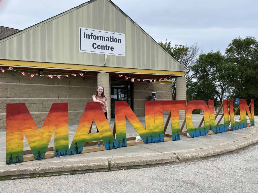 Young woman standing at manitoulin sign in little current on Manitoulin Island.
