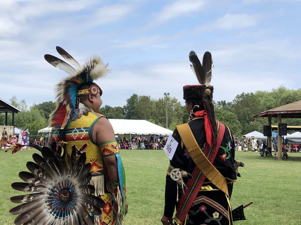 Two First Nations men dressed in regalia facing field at Wikwemikong during annual cultural festival pow wow.