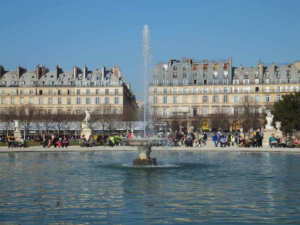 People sitting around fountain in Tuileries Gardens in Paris on spring day with buildings in background.