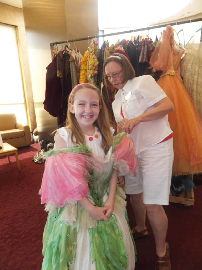 Young girl trying on pink and green costume dress with festival employee and wrap of costumes in background.