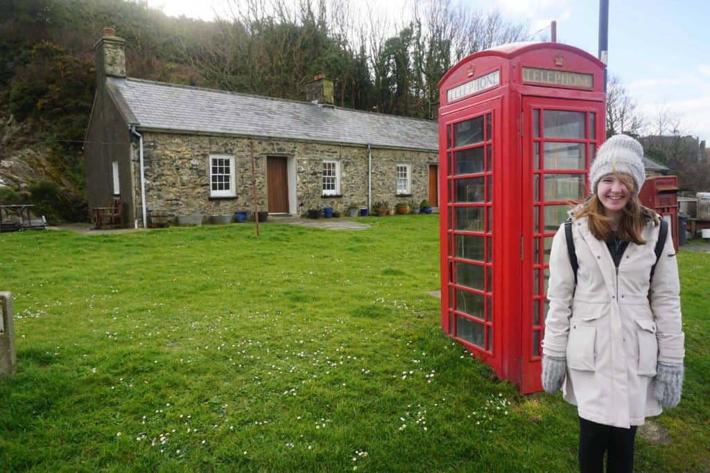 Teen girl wearing light-coloured coat, hat and mittens standing in front of red telephone box with stone building in background in Porthgain.