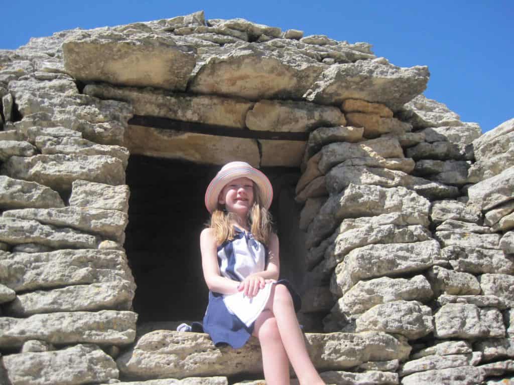 Young girl in dress and straw hat sitting with legs crossed and hands in lap in window of borie structure.
