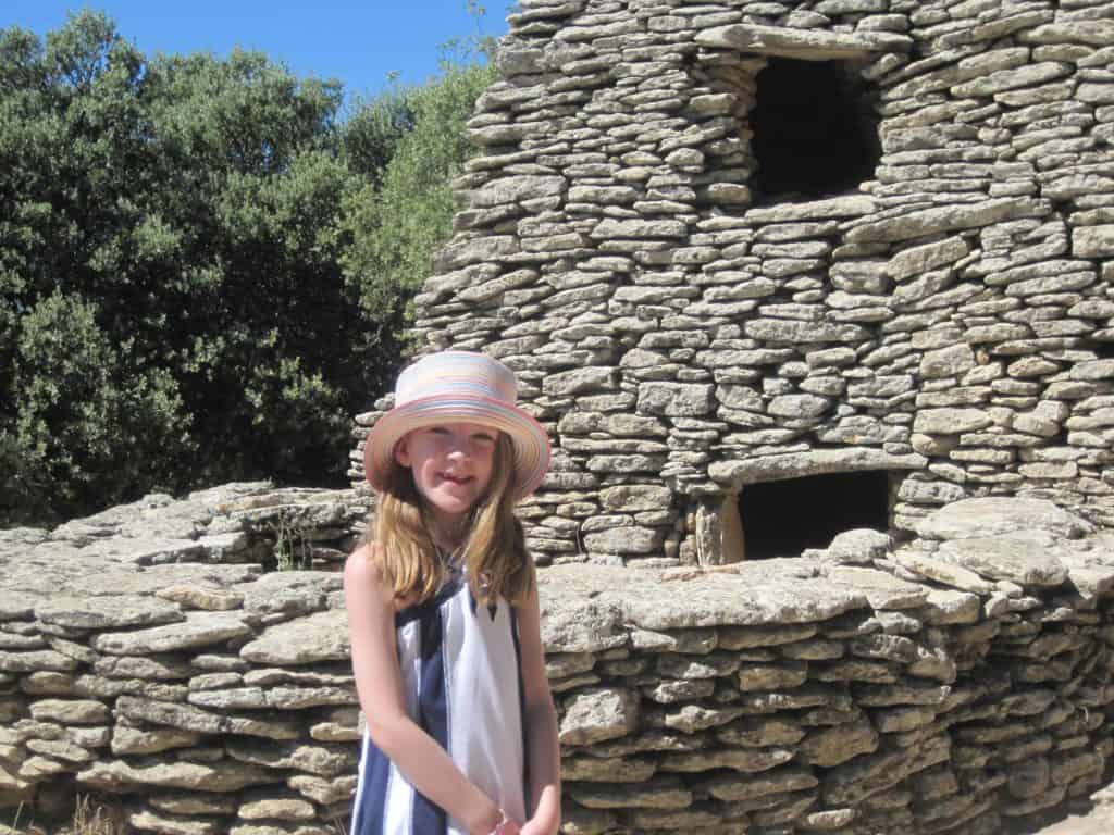 Young girl in blue and white dress and multicoloured straw hat posing in front of borie structure at Village des Bories.