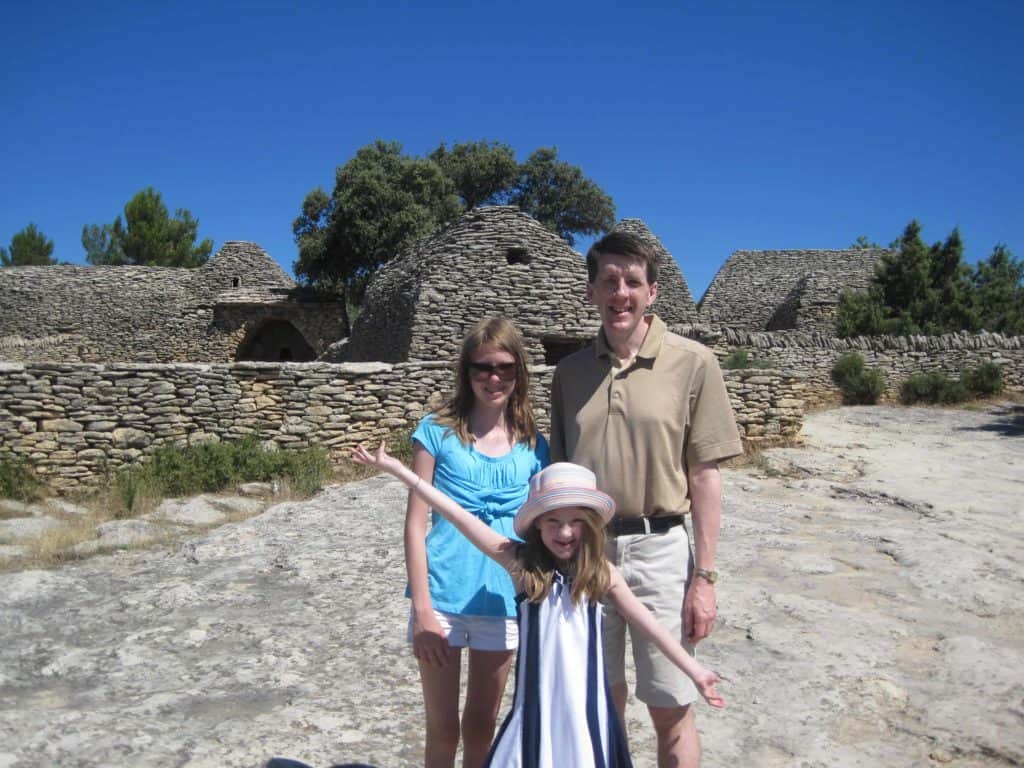 Man, teen girl and younger girl in gat with arms outstretched posing in front of borie structure at Village des Bories.