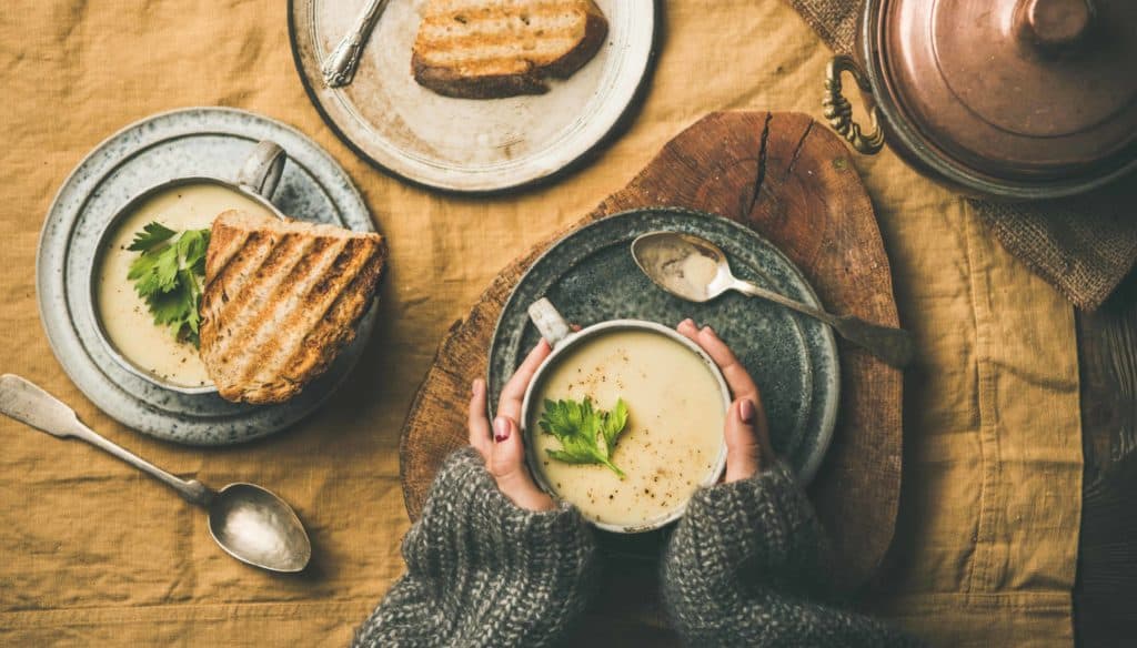 flat lay of plates of grilled cheese and a cup of soup sitting on top of a brown table cloth. Woman wearing gray sweater has hands around cup of soup.