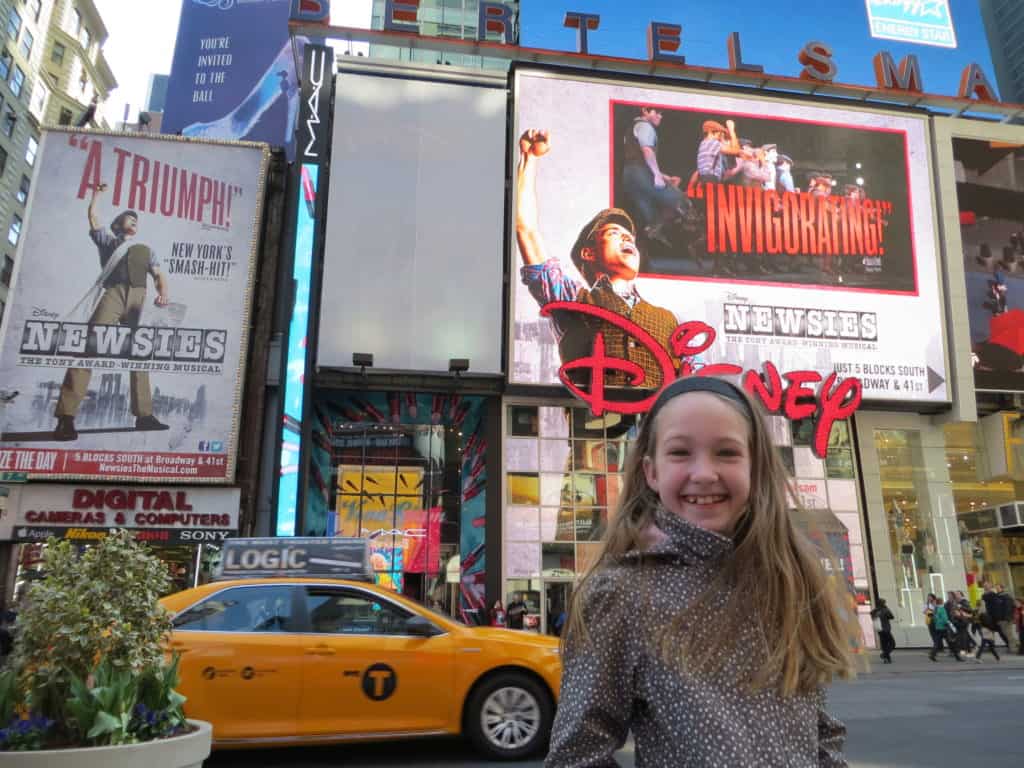Young girl standing in Times Square with billboards for Disney Newsies in background.