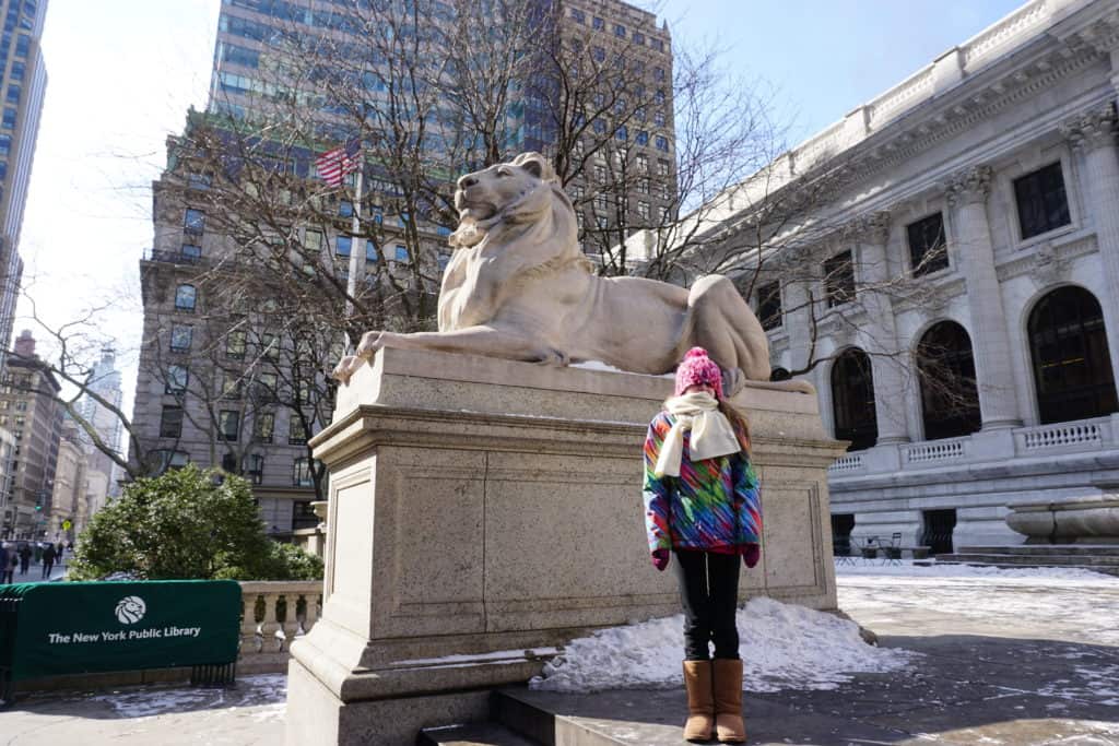 Young girl bundled up with scarf around face standing by lion sculpture outside the New York Public Library with some snow on ground.