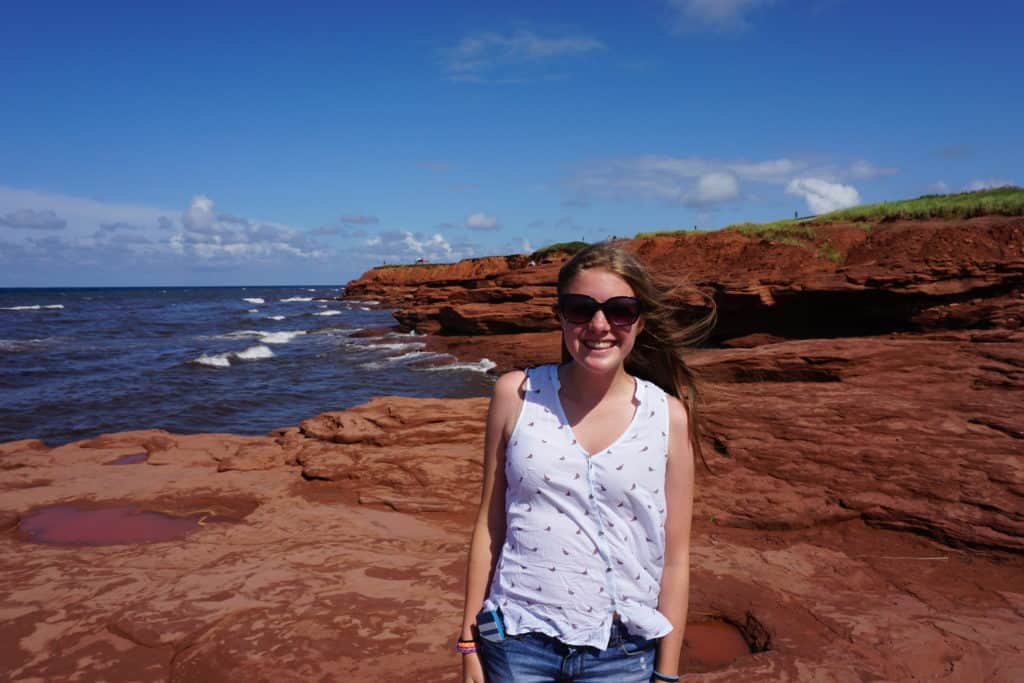 Young woman in white sleeveless top wearing sunglasses walking on red cliffs by ocean in Prince Edward Island National Park, Canada.