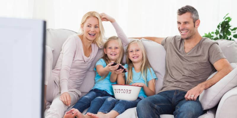 Twin girls and parents sitting on a couch watching television and eating popcorn.