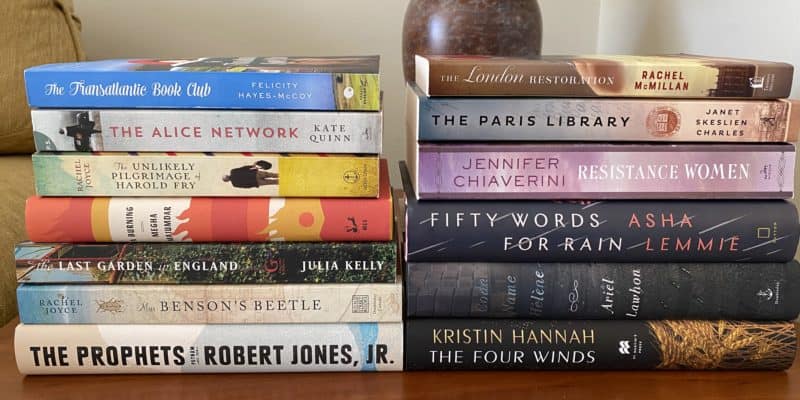 Two stacks of books on table with lamp behind.