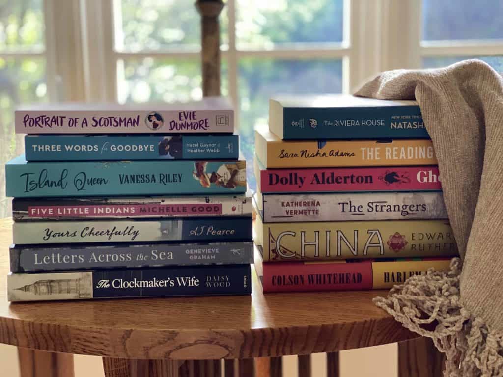 Two stacks of books sitting on a round wood table in front of a window with beige blanket draped over edge of books.