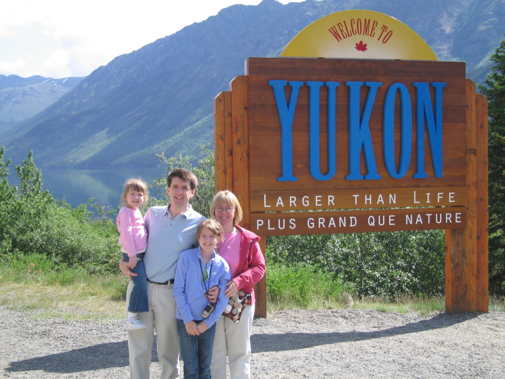 Family of four with two young girls standing at Welcome to Yukon Territory sign.