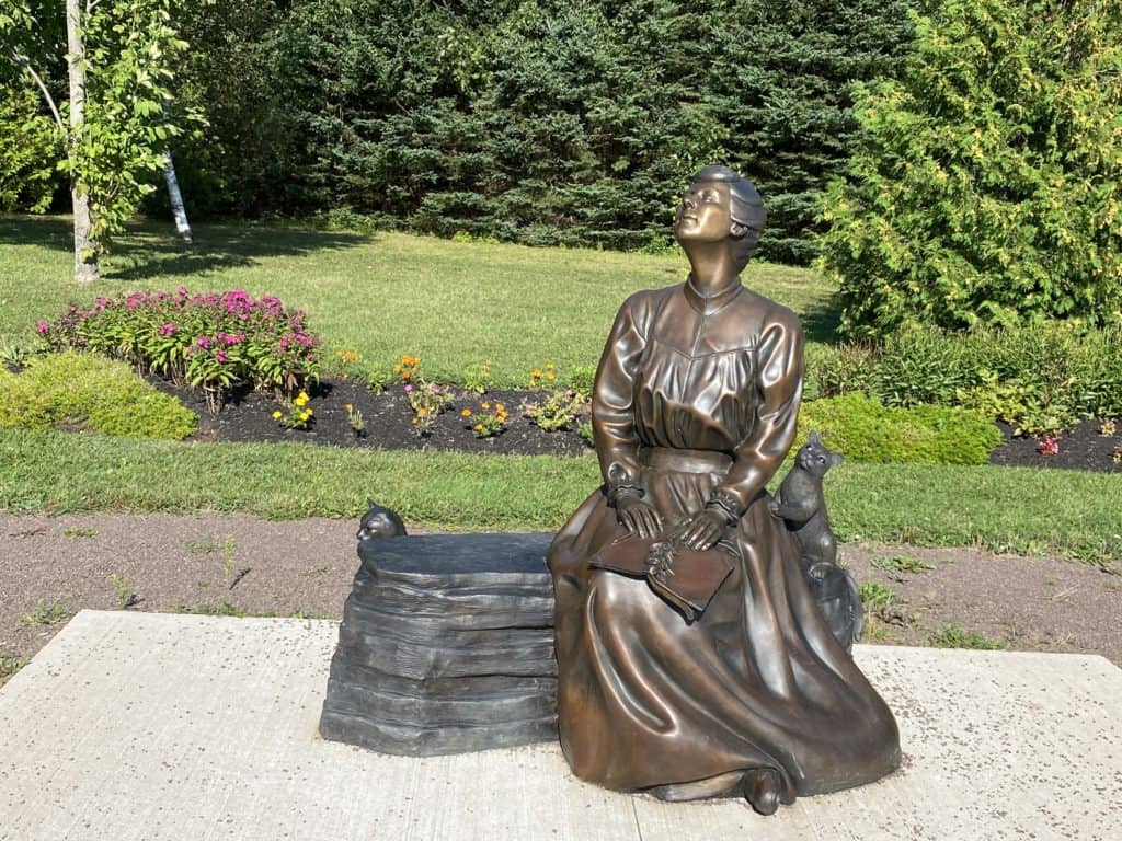 Bronze statue of Lucy Maud Montgomery sitting and holding a book with two cats in garden in Cavendish, Prince Edward Island.