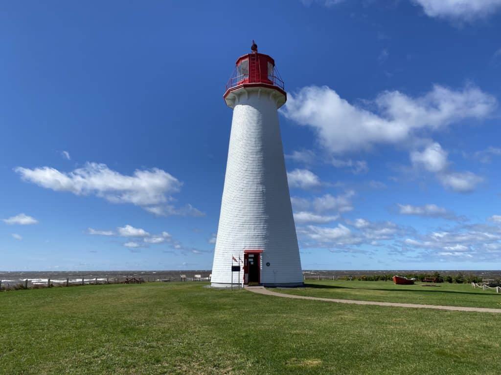 Point Prim Lighthouse, Prince Edward Island - white conical shaped lighthouse with red trim on grassy spot with ocean in background and blue skies with white wispy clouds.