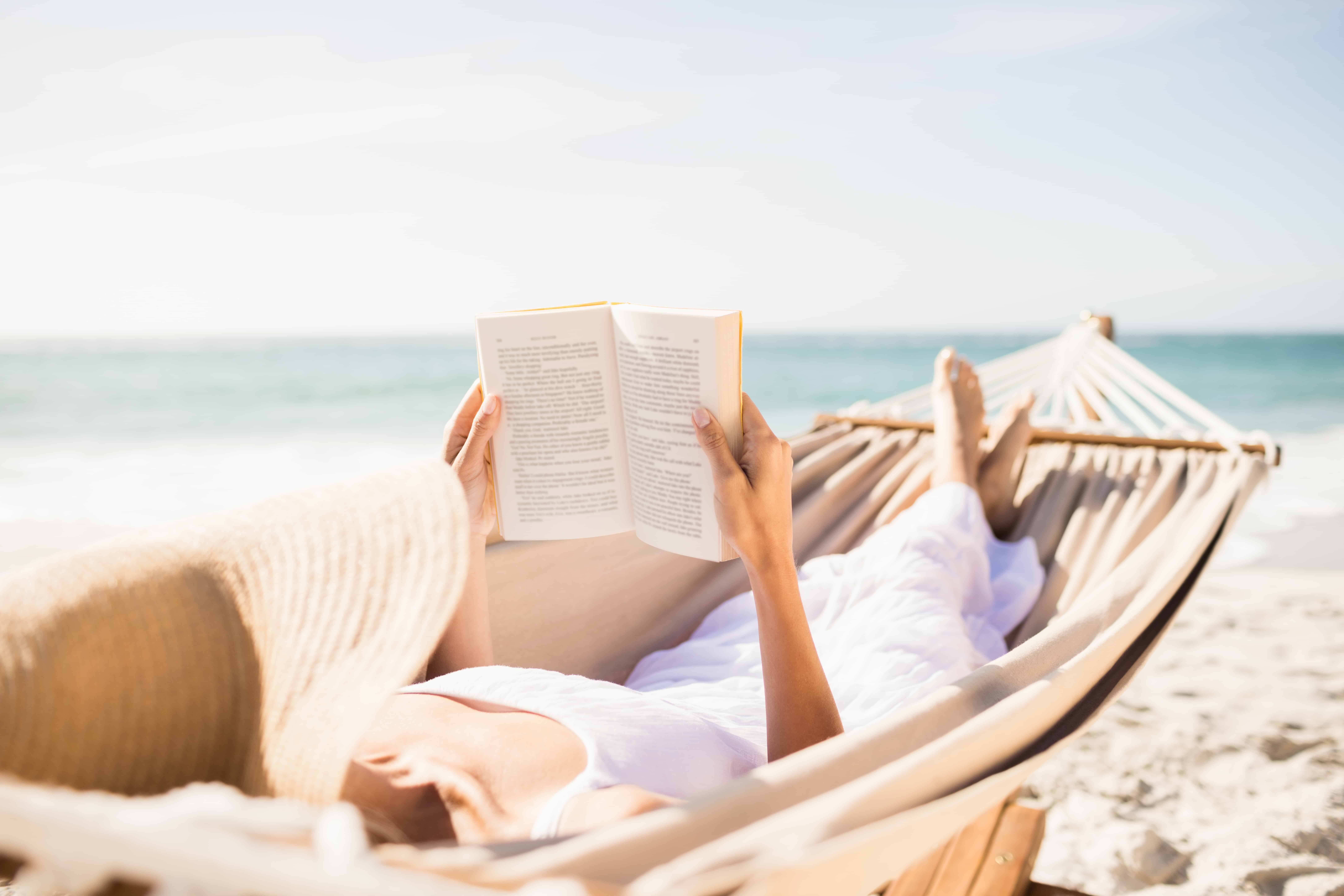 woman wearing straw sunhat reading book in hammock on the beach.