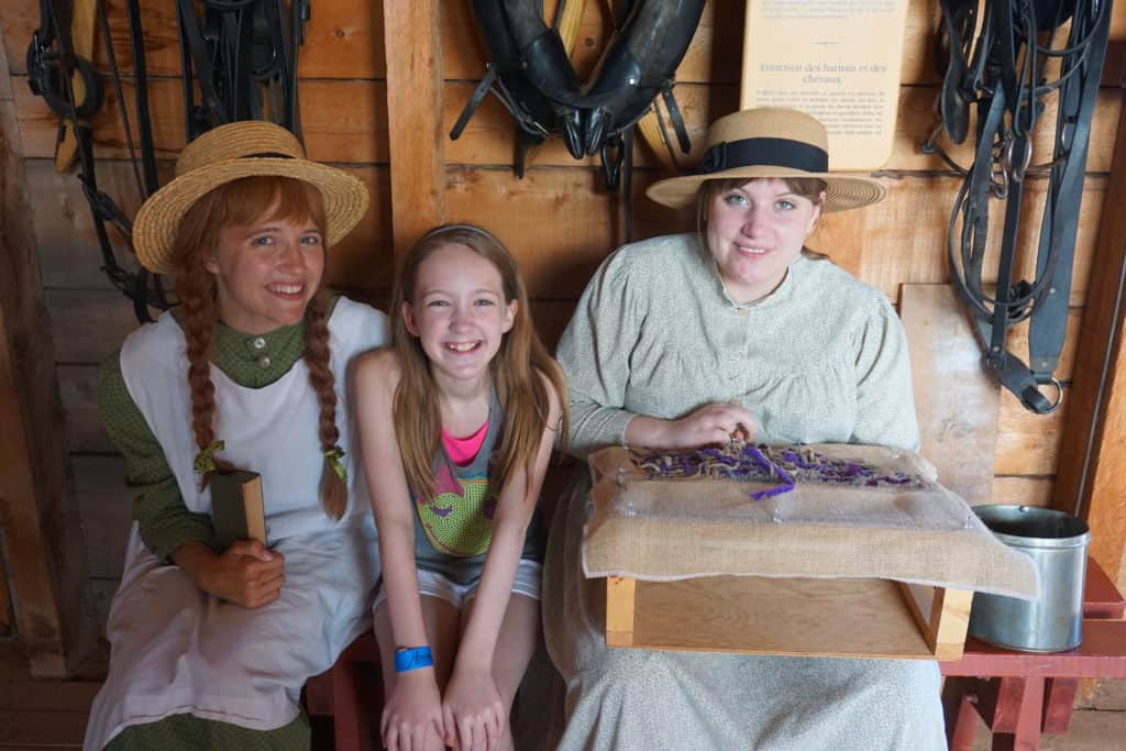 Young girl sitting between Anne of Green Gables holding book and young woman demonstrating rug hooking in barn at Green Gables, PEI.