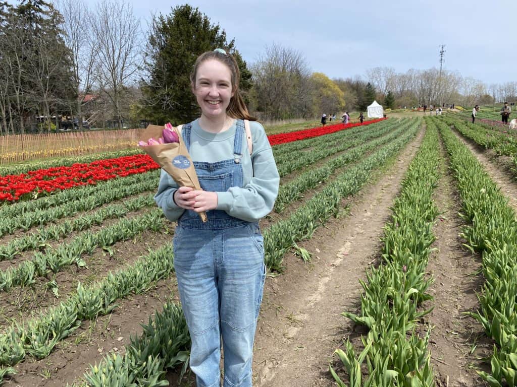 Young woman standing in front of rows of tulip plants holding bouquet of tulips wrapped in brown paper.