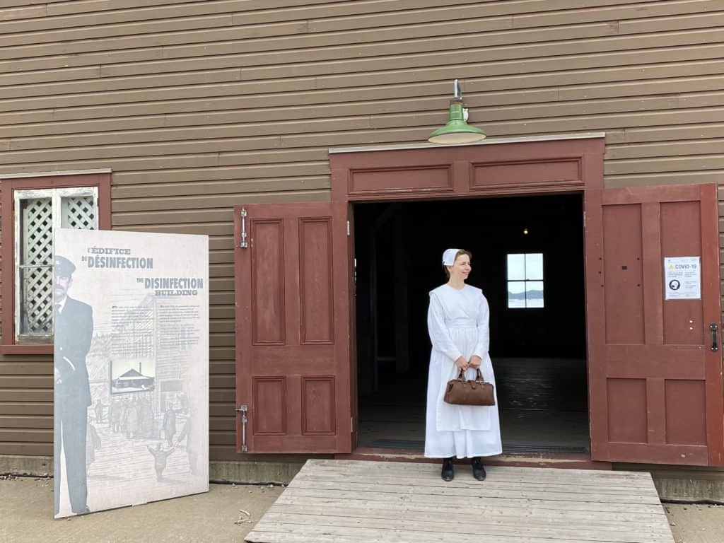 Woman in white dress, apron and hat holding brown satchel in doorway of the Disinfection Building at Grosse-Île, Québec.