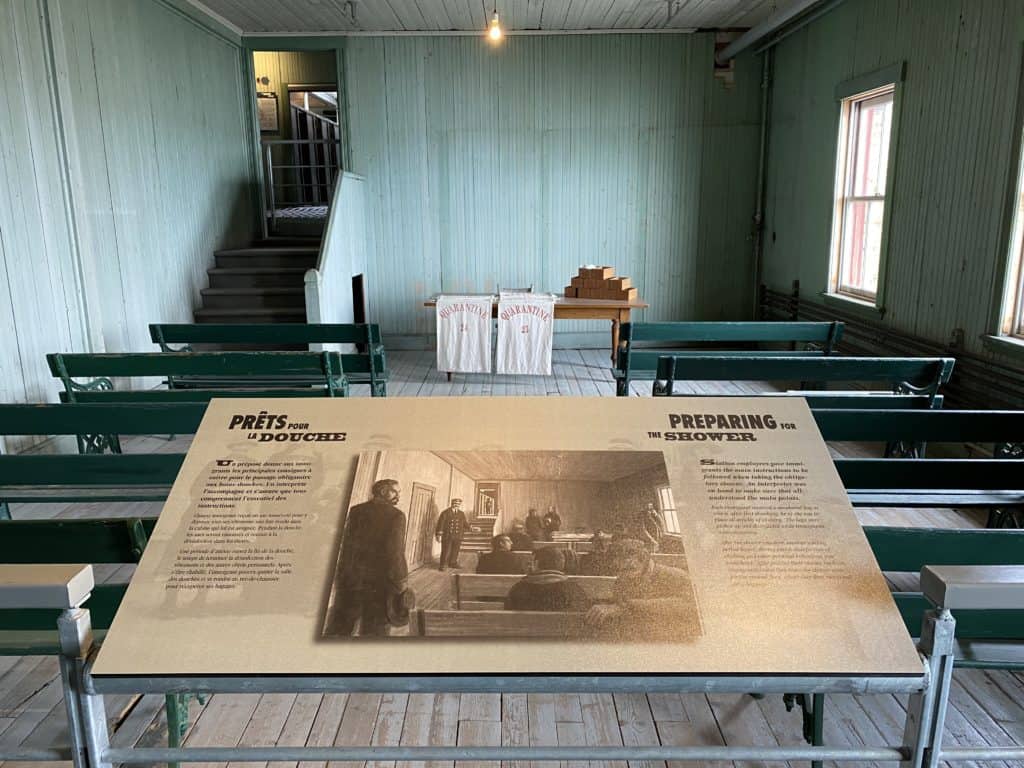 building at Grosse-Île quarantine station painted green with two rows of green benches, board with information titled Preparing for the Shower.