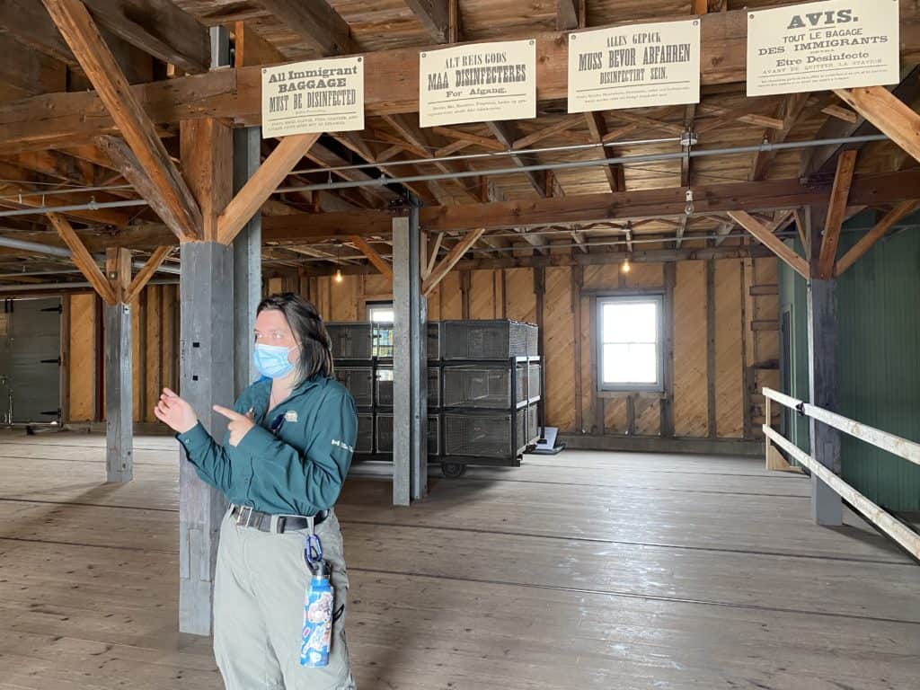 Female Parks Canada guide wearing blue surgical mask standing inside building explaining the disinfection process for immigrants arriving at the quarantine station at Grosse-Ile, Quebec