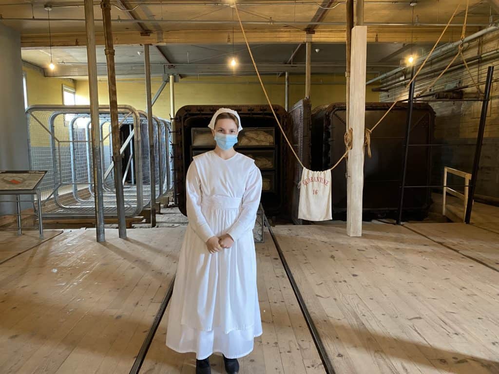 Woman dressed as nurse in white dress, apron and cap with blue surgical mask inside medical inspection bulding at Grosse-Ïle, Québec.