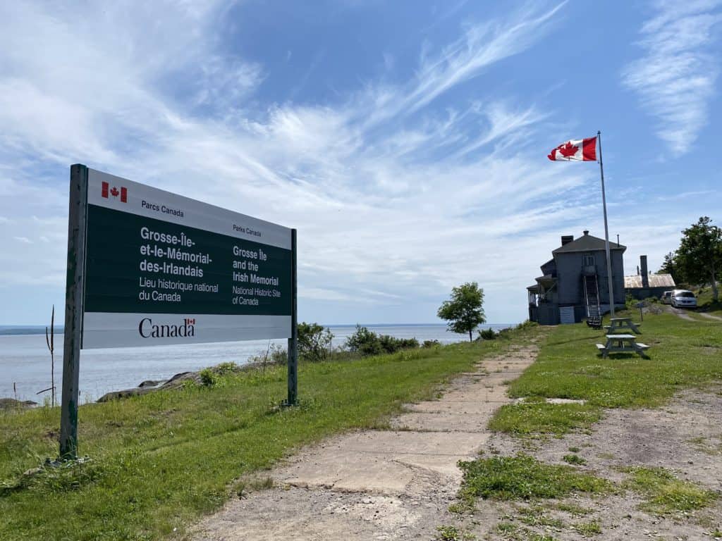 Arriving at Grosse-Ile National Historic Site of Canada - Parks Canada sign, Canada flag, picnic tables and historic building along shore of St. Lawrence.