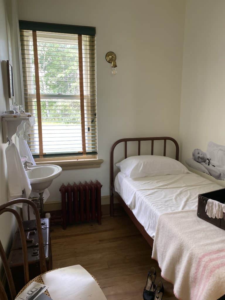 Room at hotel at Grosse-Île, Québec quarantine station with small bed with pillow, pink striped wool blanket, sink, towels hanging and shelf over sink, chair and leather suitcase.