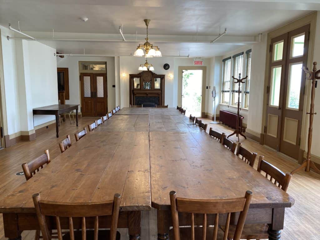 Room with long wooden table and chairs with chandeliers overhead and fireplace at end of room.