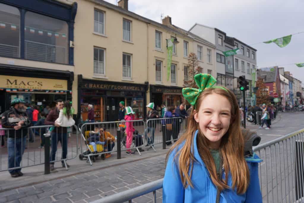 Smiling girl in blue coat wearing green shamrock headband on street in Killarney waiting for start of St. Patrick's Day Parade.