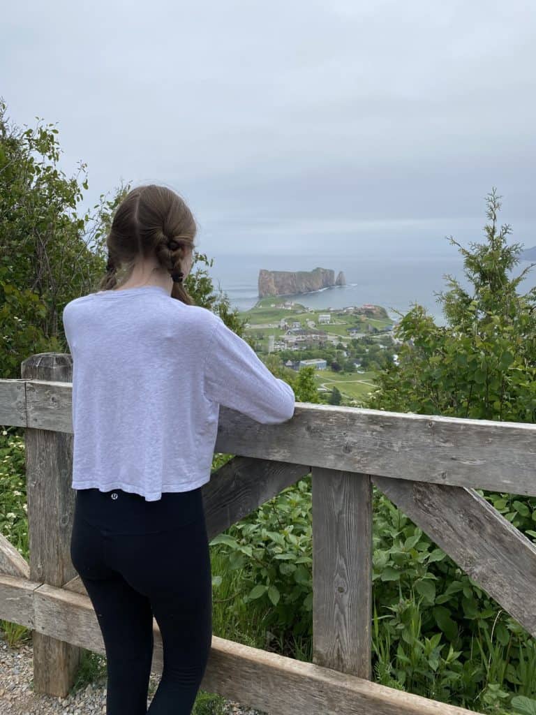 Young woman in breads standing at wooden fence looking at Perce Rock and village.