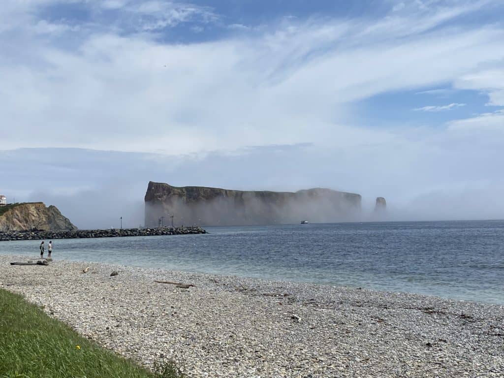 Rocky beach by ocean with Perce Rock shrouded in mist and a bit of blue sky peeking through.