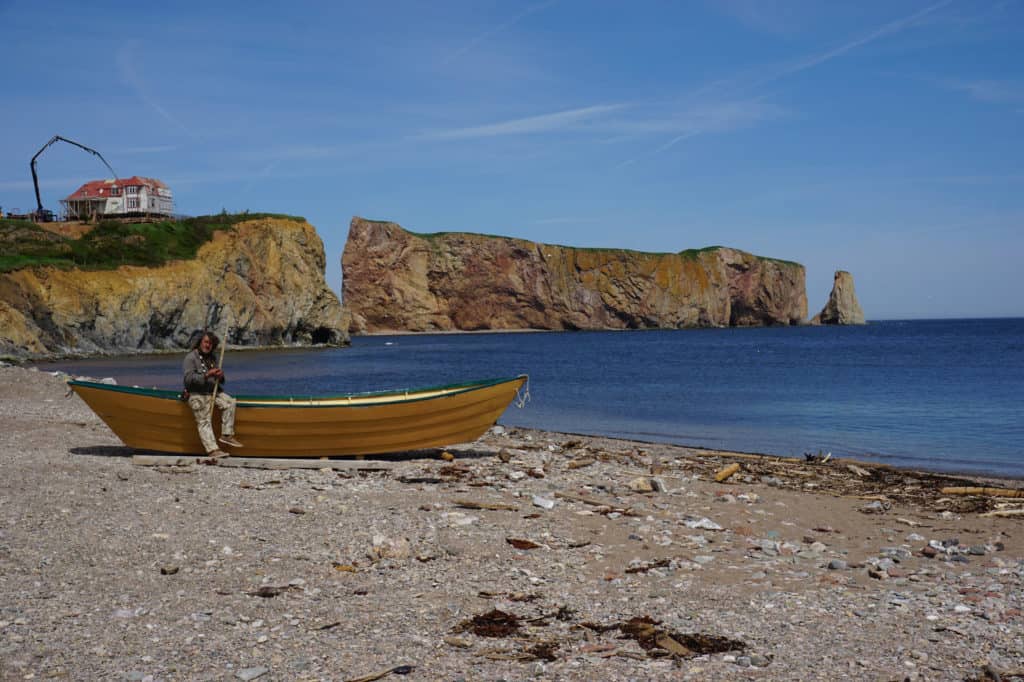 Man sitting on yellow row boat on sand beach with Perce Rock in background.