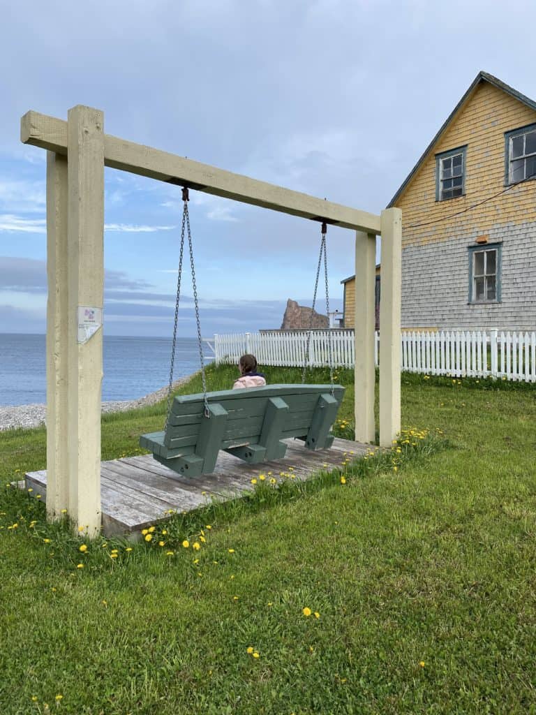 Young woman sitting on green bench swing beside clapboard building and white picket fence by ocean with Perce Rock partially visible in distance.