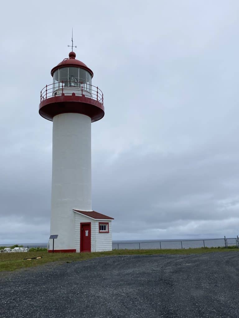 Cap-de-la-Madeleine lighthouse - white cylindrical tower topped with red light on overcast day with water in background.