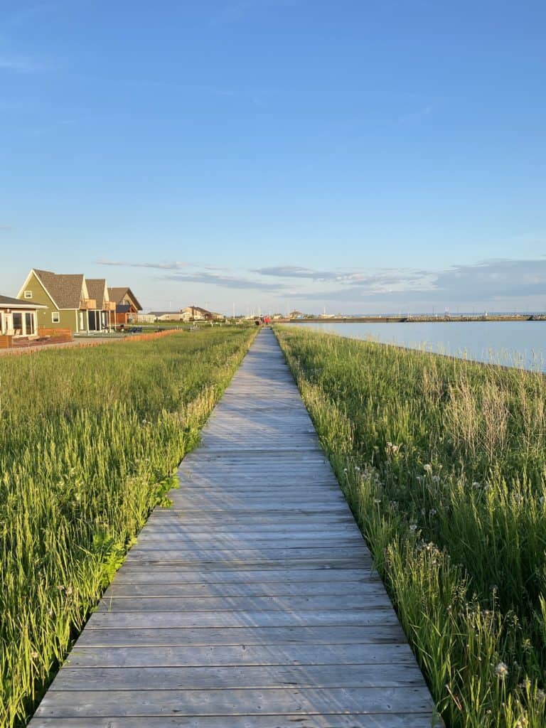 Wooden boardwalk running through grassy area with small wooden buildings on one side and water on the other in Carleton-sur-Mer, Quebec.