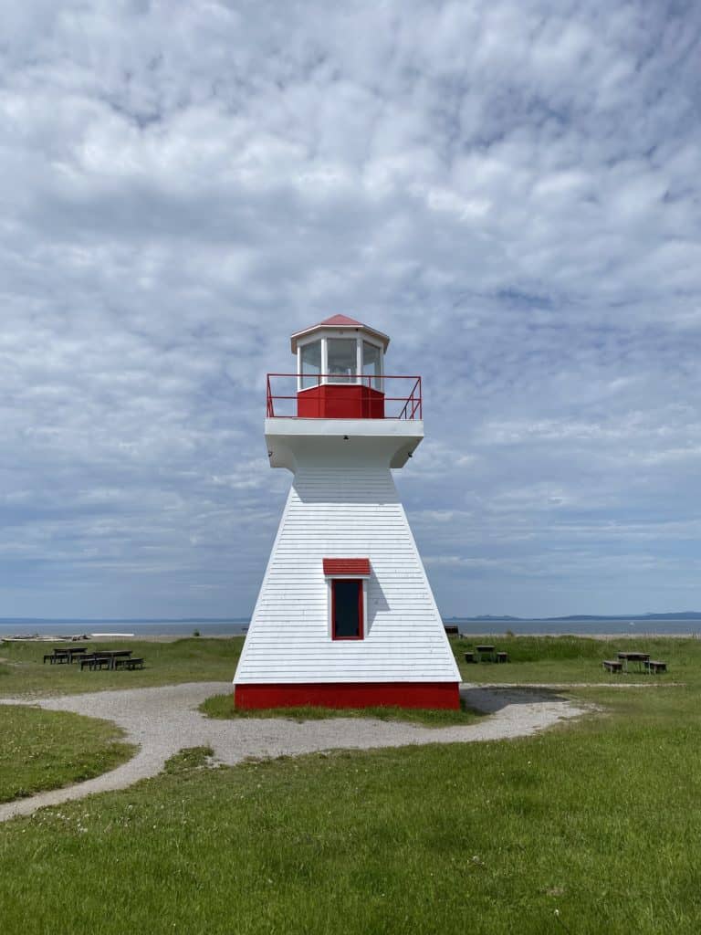 Small white lighthouse with red trim surrounded by picnic tables with water in the background in Carleton-sur-Mer, Quebec.