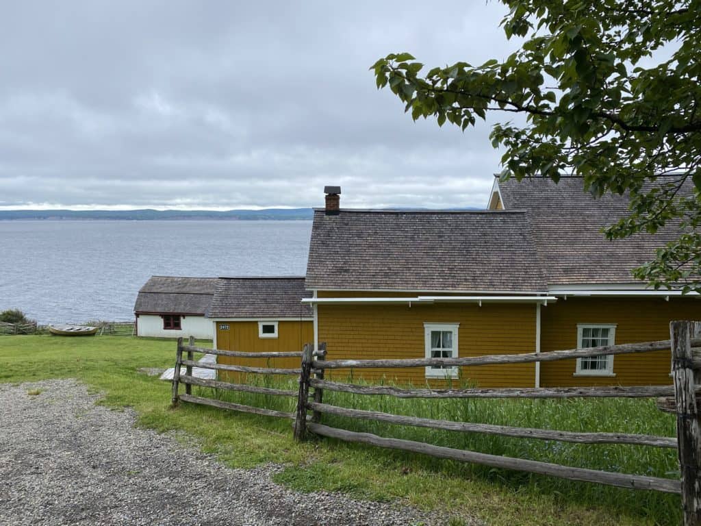 historic wooden buildings with dark yellow siding, white windows and grey shingled roof with fence in foreground and water in background in Forillon National Park.