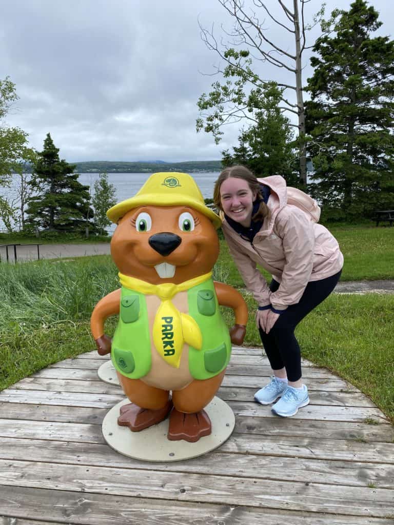 young woman in black pants and pink windbreaker jacket crouched beside Parks Canada beaver mascot statue wearing green vest and yellow tie and hat.