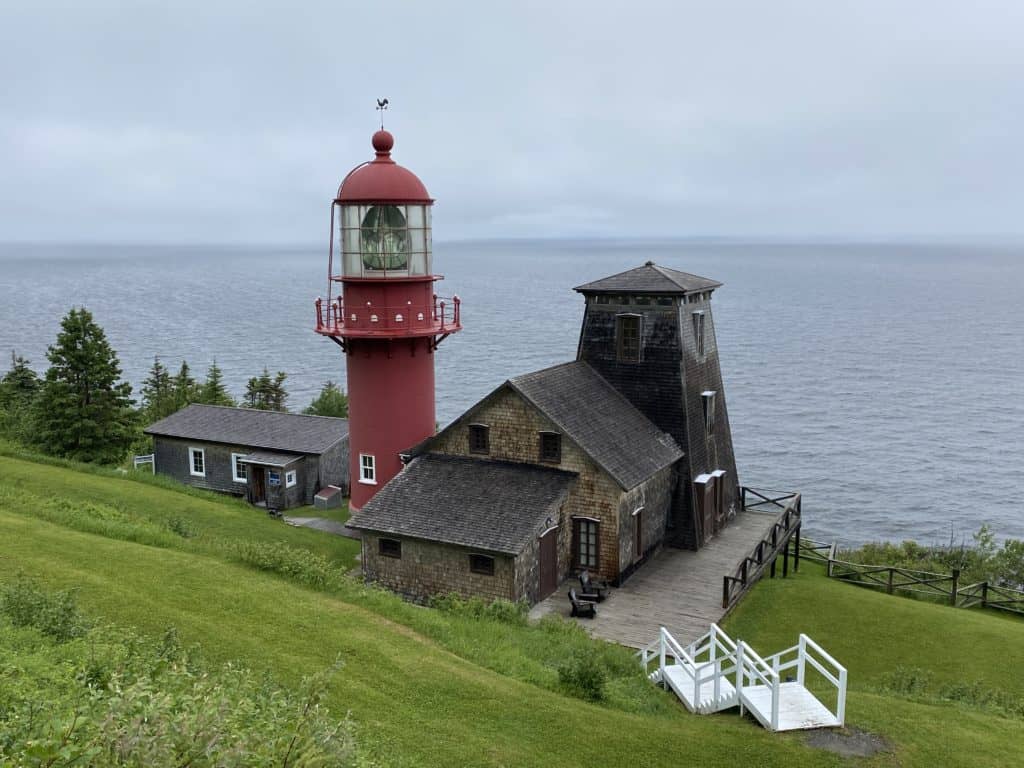 bright red cylindrical lighthouse with brown keepers buildings on both sides sits on green grassy hill with water in background on over cast day in Gaspe region of Quebec.